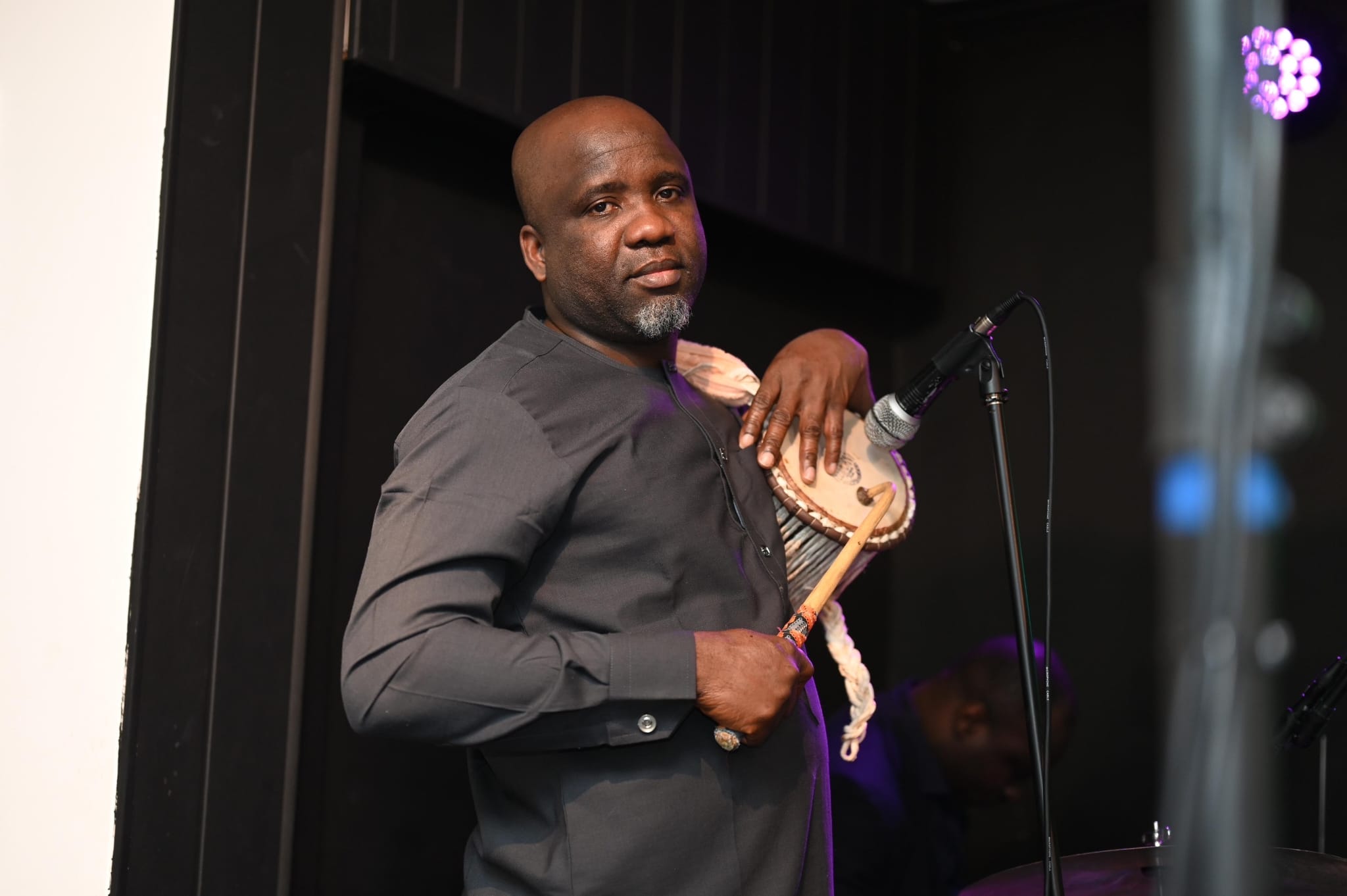 Ipadeola Sunday Abidemi playing talking drum with curved stick in purple stage light